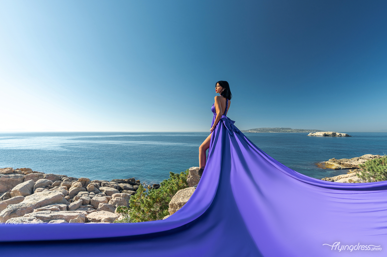 A woman in a flowing purple dress stands on a rocky cliff overlooking the expansive blue sea and distant islands of Kavouri, Athens.